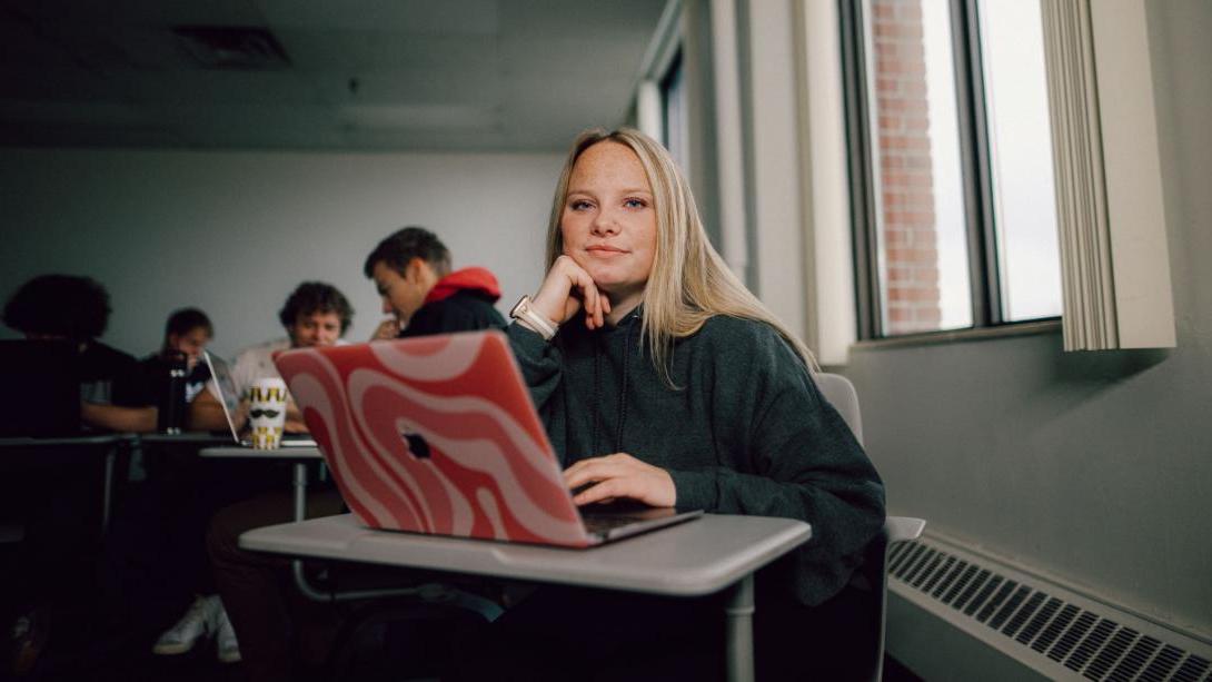 A Kettering management student sits in a classroom. On the desk is a laptop with a pink case.